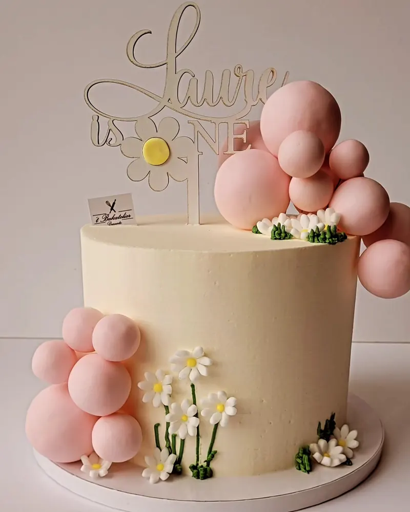 Crèmekleurige taart met roze suikerballen, madeliefjes en een naamtopper